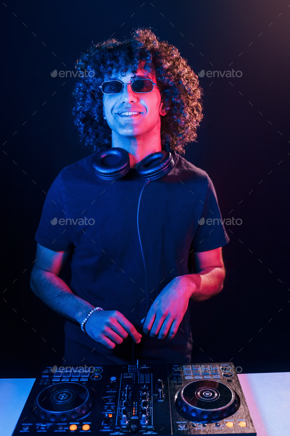 Man with curly hair using DJ equipment and standing in the dark neon lighted room