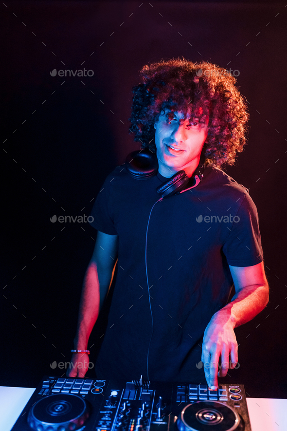 Man with curly hair using DJ equipment and standing in the dark neon lighted room