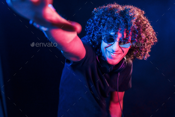 Young DJ with curly hair standing in the club with neon light