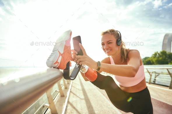 Staying in touch. Happy disabled woman in sportswear and headphones stretching prosthetic leg and