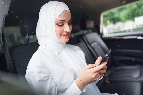 Beautiful muslim woman wearing white hijab sitting on the back seat of a car and using smartphone