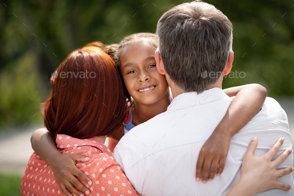Dark-skinned appealing cute girl hugging foster parents