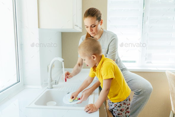 mom and her toddler son wash dishes in the kitchen sink. The child helps to do household chores.