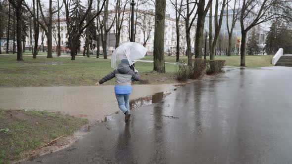 Boy with Transparent Umbrella Runs Along Wet Pavement Roads in City Park alt