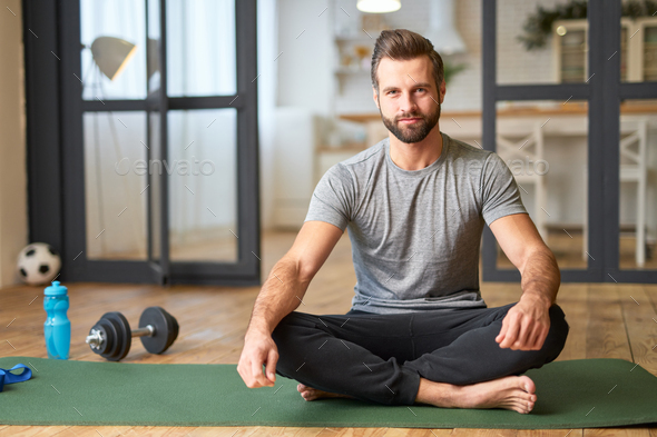 Handsome young man sitting on yoga mat at home Stock Photo by friends_stock