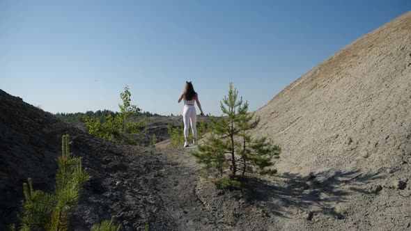 A Girl in a Sporty Uniform Walks Along a Sandy Mountain Road Towards the Lake alt