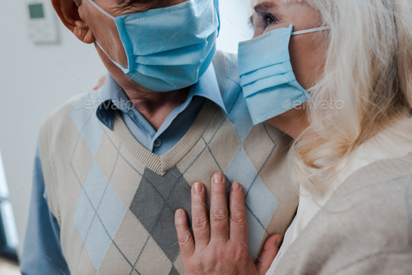cropped view of elderly couple in medical masks hugging at home during self isolation