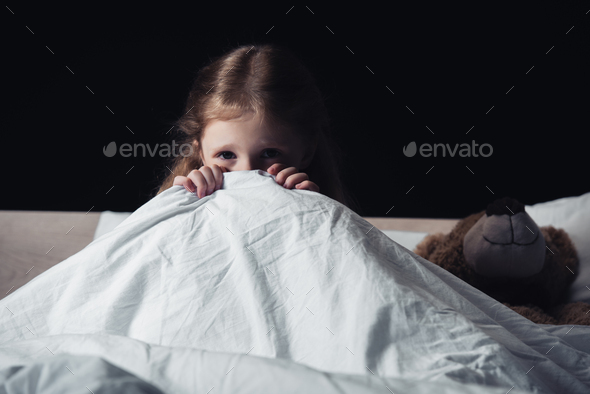 scared child hiding under blanket while sitting on bedding near teddy bear isolated on black