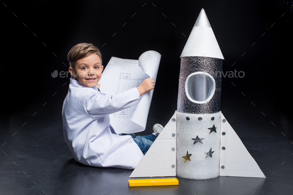 Little boy in white coat holding blueprint while sitting near toy rocket and smiling at camera