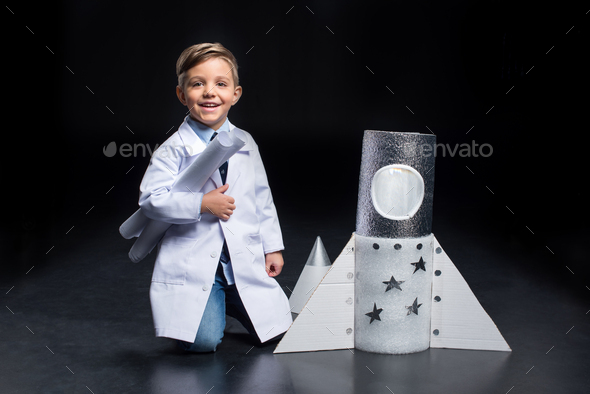 Smiling little boy in white coat holding blueprints near toy rocket