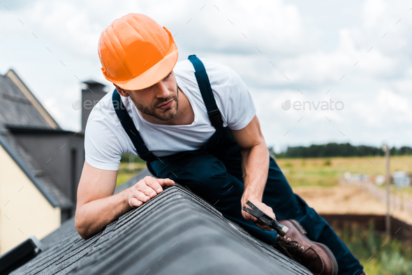 selective focus of repairman in orange helmet sitting on roof and holding hammer