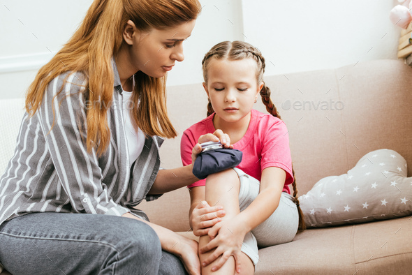 worried mother holding ice bag compress on painful knee of daughter
