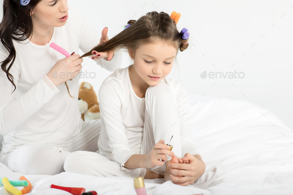 Mother curling hair to daughter applying nail polish on toenails isolated on white
