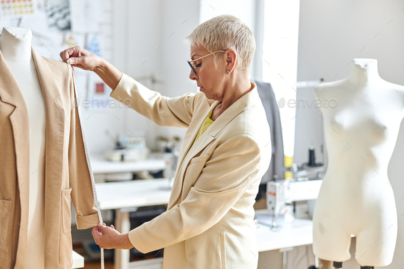 Middle aged woman designer measures sleeve of elegant jacket in light workshop