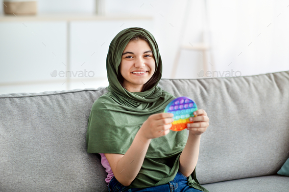 Smiling Indian teen girl in hijab sitting on comfy couch, playing with trendy POP IT toy at home