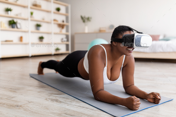Domestic virtual training. Curvy young black lady in VR glasses standing elbow plank at home