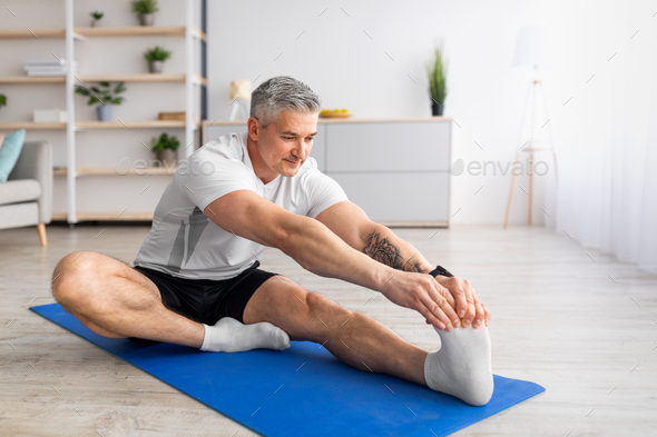 Home workout. Sporty mature man stretching her leg on yoga mat in living  room, free space Stock Photo by Prostock-studio