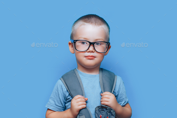pretty blonde boy on blue background with backpack unhappy, sad, child don\'t want go to school