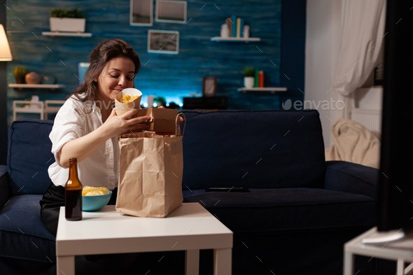 Smiling cheerful woman unpacking fast food meal delivery sitting on sofa during junk-food home