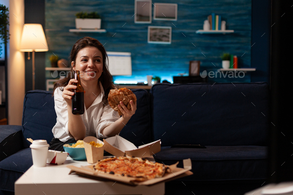 Happy woman holding beer bottle eating tasty takeaway food delivery while watching entertainment