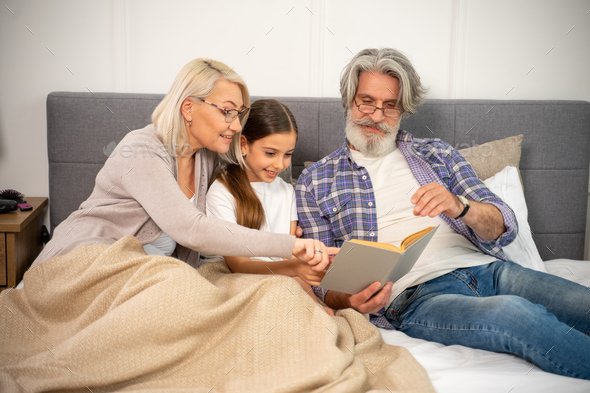 Smiling senior grandparents reading book with little girl lying on bed