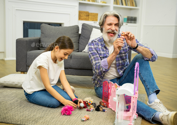 Smiling grandfather having fun and playing with granddaughter showing him dollhouse