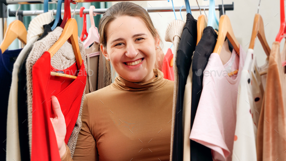 Beautiful smiling woman behind long rack of clothes on hangers choosing dress to wear