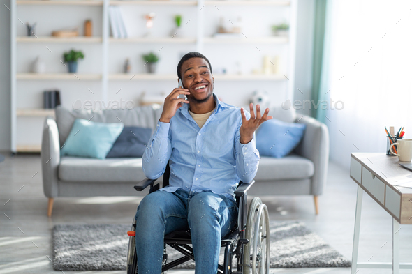 Paraplegic black guy in wheelchair speaking on smartphone with friend at home