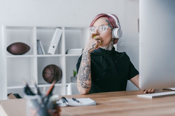 young tattooed businesswoman in headphones looking away at workplace