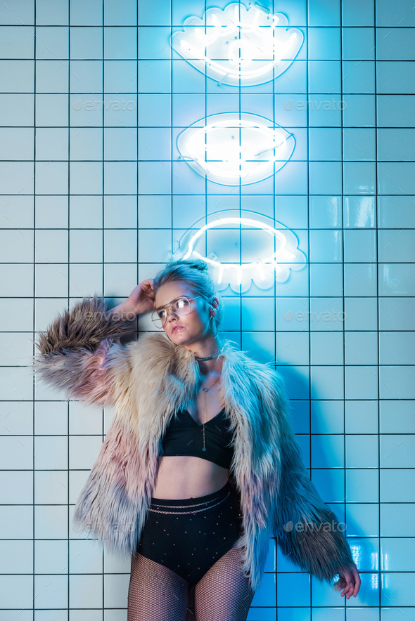 attractive fashionable young woman posing in black sexy clothes and fur coat near neon signs
