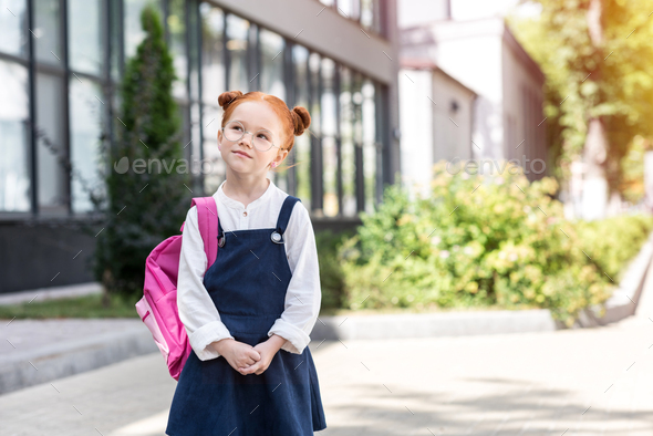 pensive red haired schoolgirl in spectacles wearing backpack and looking away - Stock Photo - Images