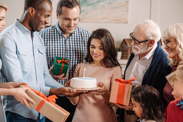 large family presenting cake and gifts to beautiful surprised woman