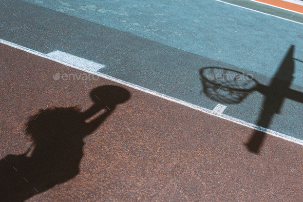 Shadows of female figure throwing ball into basket on ground of sports field