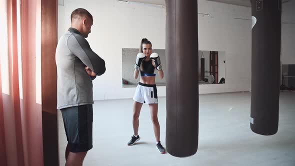 A Strict Trainer Watches His Female Kickboxer Student Practice a Punch on a Punching Bag in a alt
