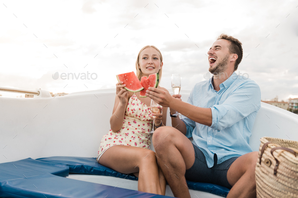 Happy young couple cheering with champagne and eating fruits on sail boat in summer vacation - Stock Photo - Images