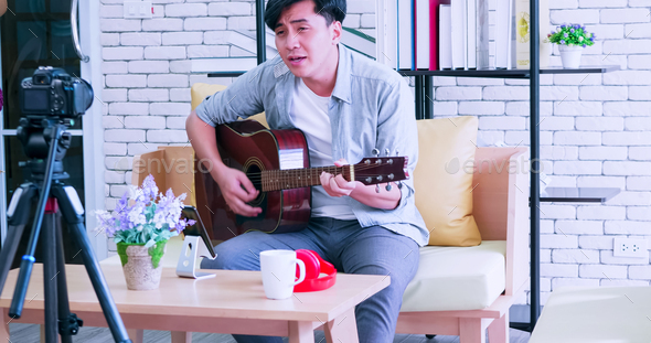 A young Asian man is playing the guitar and singing on social media by streaming live from his home.