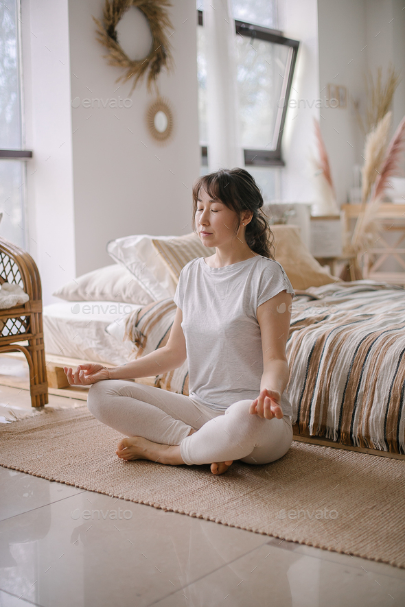 Asian woman sitting on floor in lotus position in bedroom. Concept of meditation, home yoga wellness
