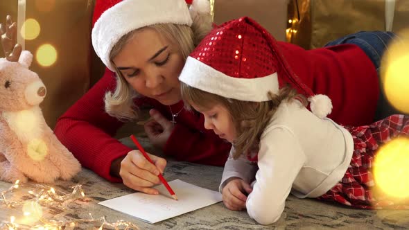 Mother and Little Child Girl Write Letter to Santa Claus on Background of Boxes with Gifts