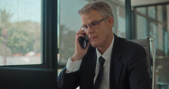 Close up of a businessman in formal wear and glasses answering a phone while working on a laptop  alt