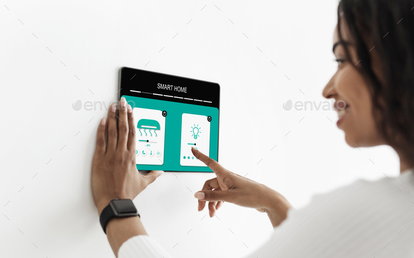 Woman controlling smart home with digital touch screen panel installed on white wall in living room