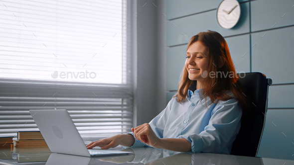 Smiling worker brunette with long loose hair types on grey laptop