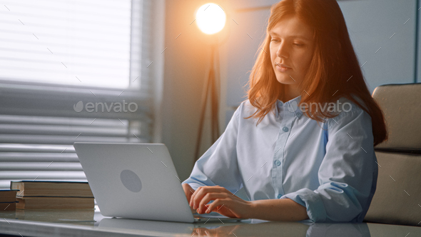 Concentrated red haired nurse with long loose hair types on grey laptop