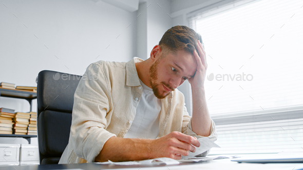 Young man bank accountant with beard looks at paper checks and puts hand on head