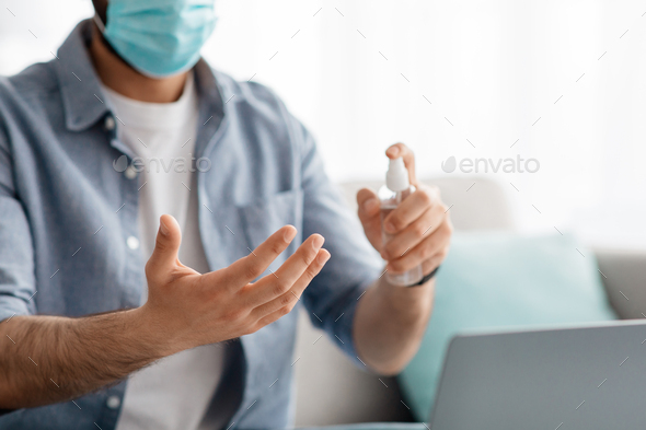 Unrecognizable man in face mask disinfecting hands with disinfectant mist spray while using modern