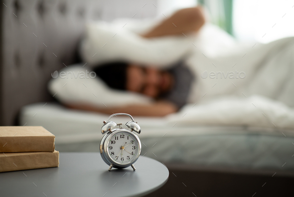 Bad sleep concept. Exhausted eastern man being awaken with alarm clock in his bedroom, covering ears