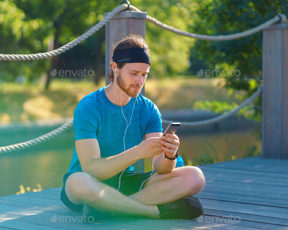 Calm young sportive man in earphones using modern smartphone while resting after exercising in park - Stock Photo - Images