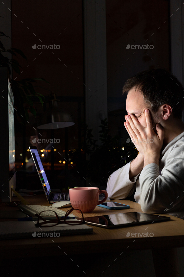Tired freelancer man rubbing eyes, working at computer at night, falls asleep from fatigue.