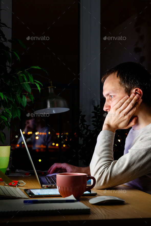 Bored man working on computer at night, can not sleep, sharing social media. Insomnia. Nomophobia.