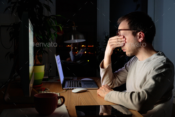 Tired freelancer man rubbing eyes, working at computer at night, falls asleep from fatigue.