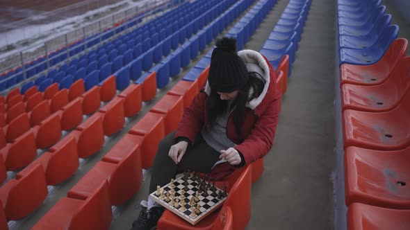 Young Woman in Winter Clothes Plays Chess Sitting on Stadium Bleachers Alone alt